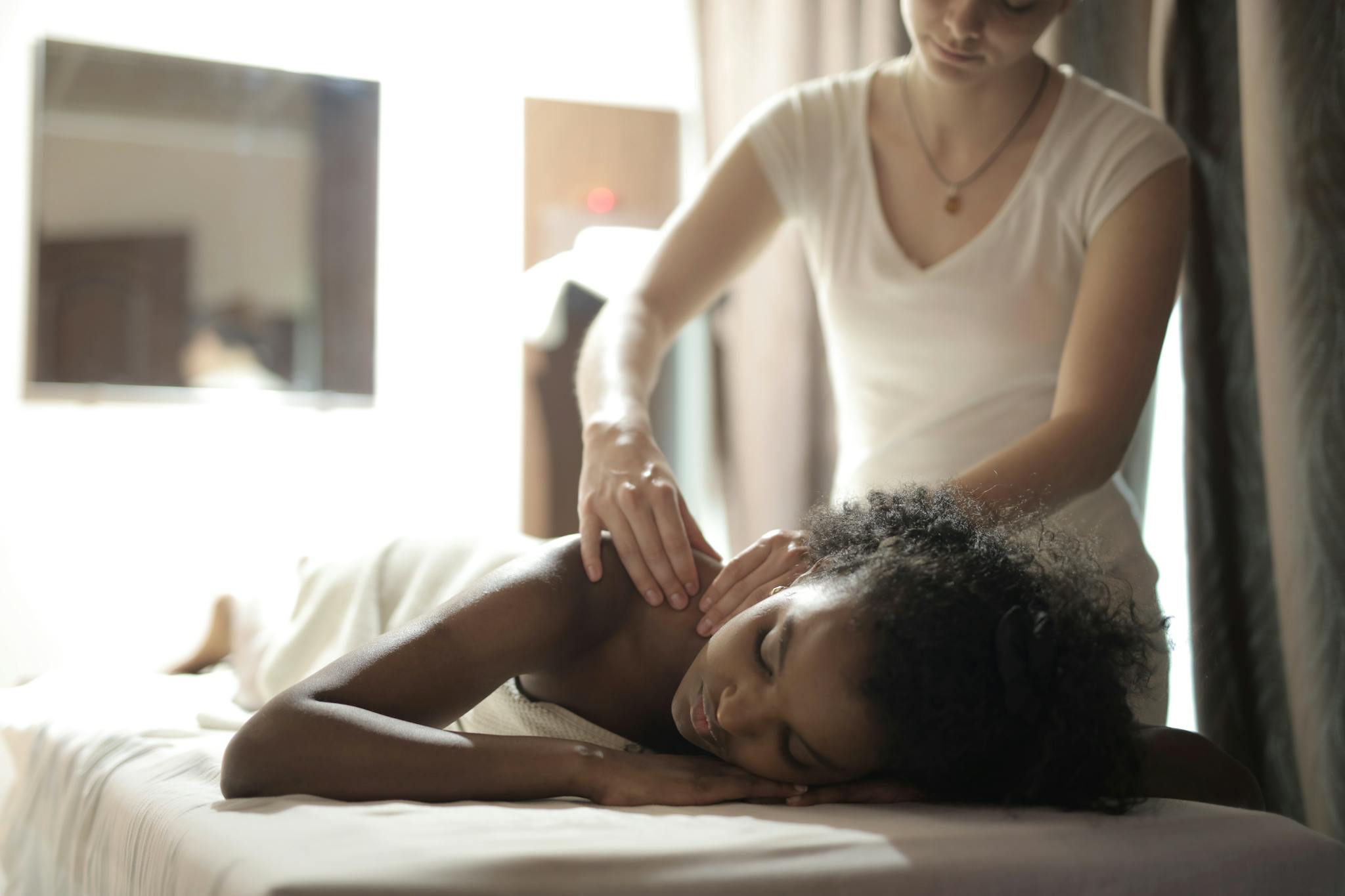 Serious young masseuse in white uniform doing massage of shoulder of young ethnic female client in light modern salon